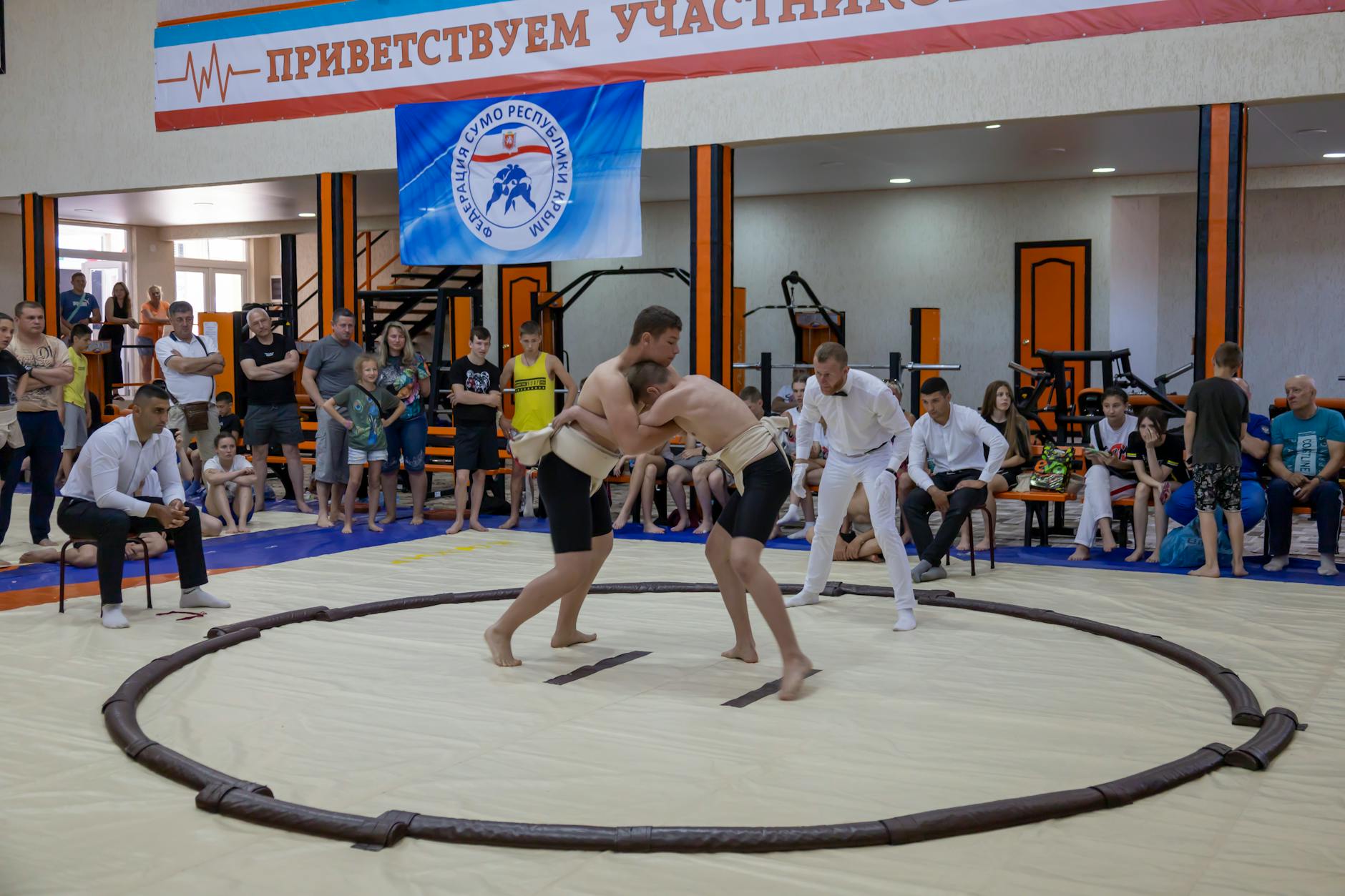 Young athletes engaged in sumo wrestling in a gym with spectators and a referee observing.