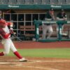 Baseball player swinging at a pitch during a game with coaches watching from the dugout.