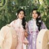 Two women in traditional Asian attire with floral elements, holding parasols outdoors.
