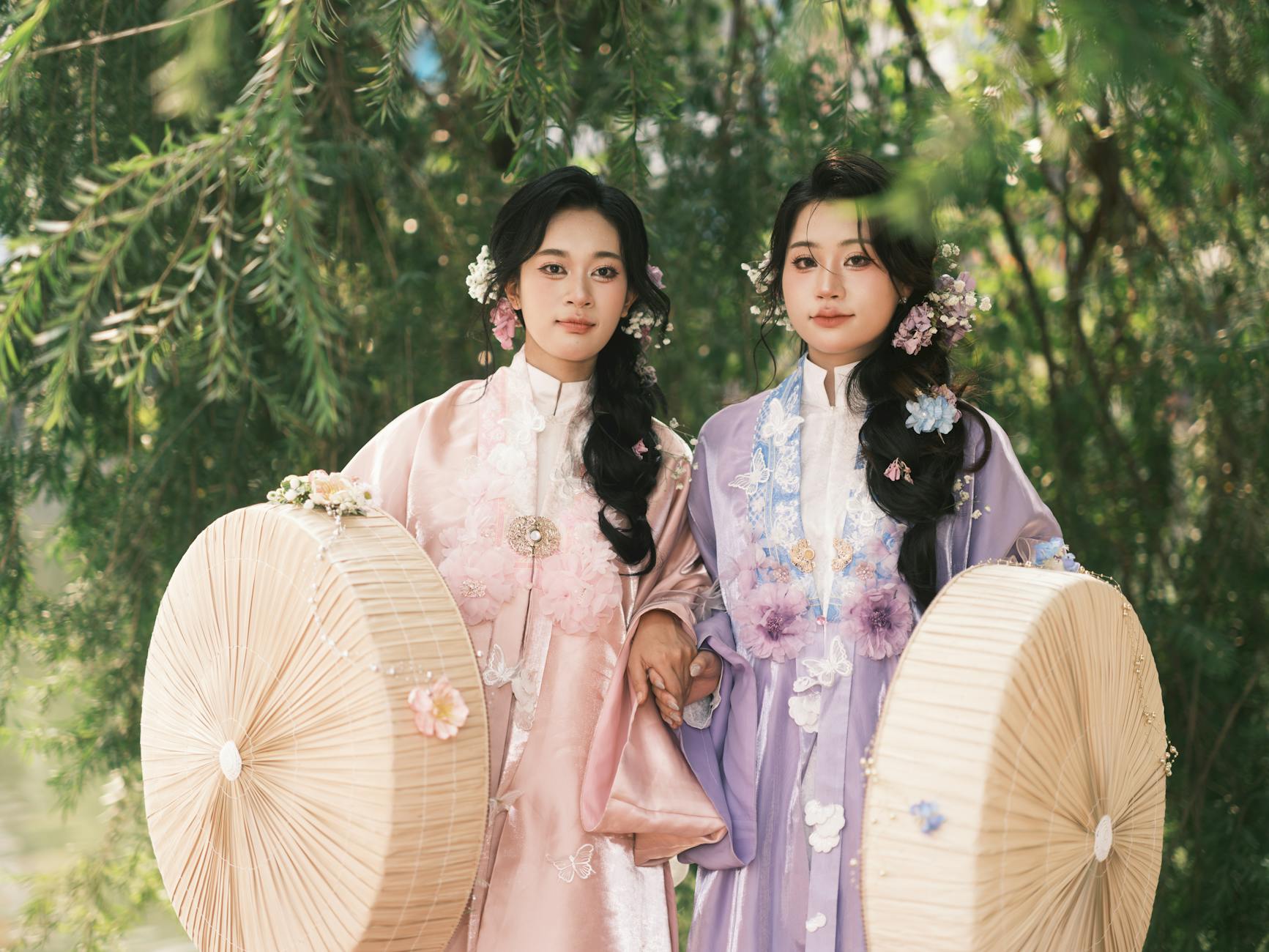 Two women in traditional Asian attire with floral elements, holding parasols outdoors.