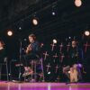 Musical performance featuring guitar and cajon onstage with performers under stage lights.