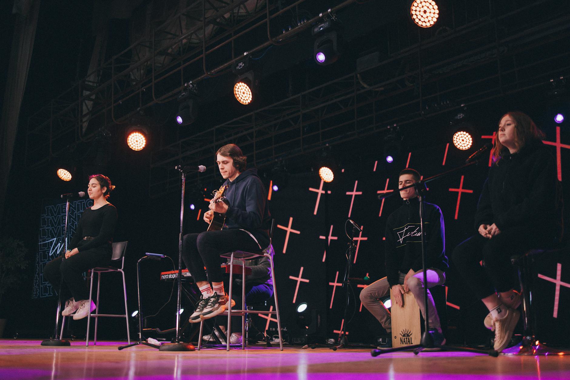 Musical performance featuring guitar and cajon onstage with performers under stage lights.