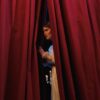 Woman peeks through red theater curtains, capturing a theatrical moment.