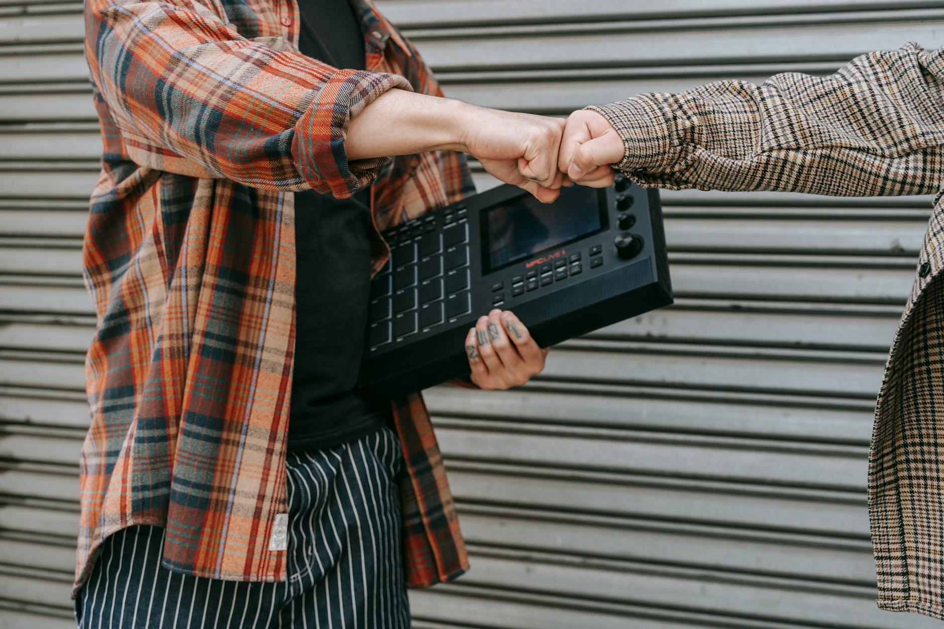 Two people in plaid shirts exchanging a fist bump while holding music equipment against a metal backdrop.