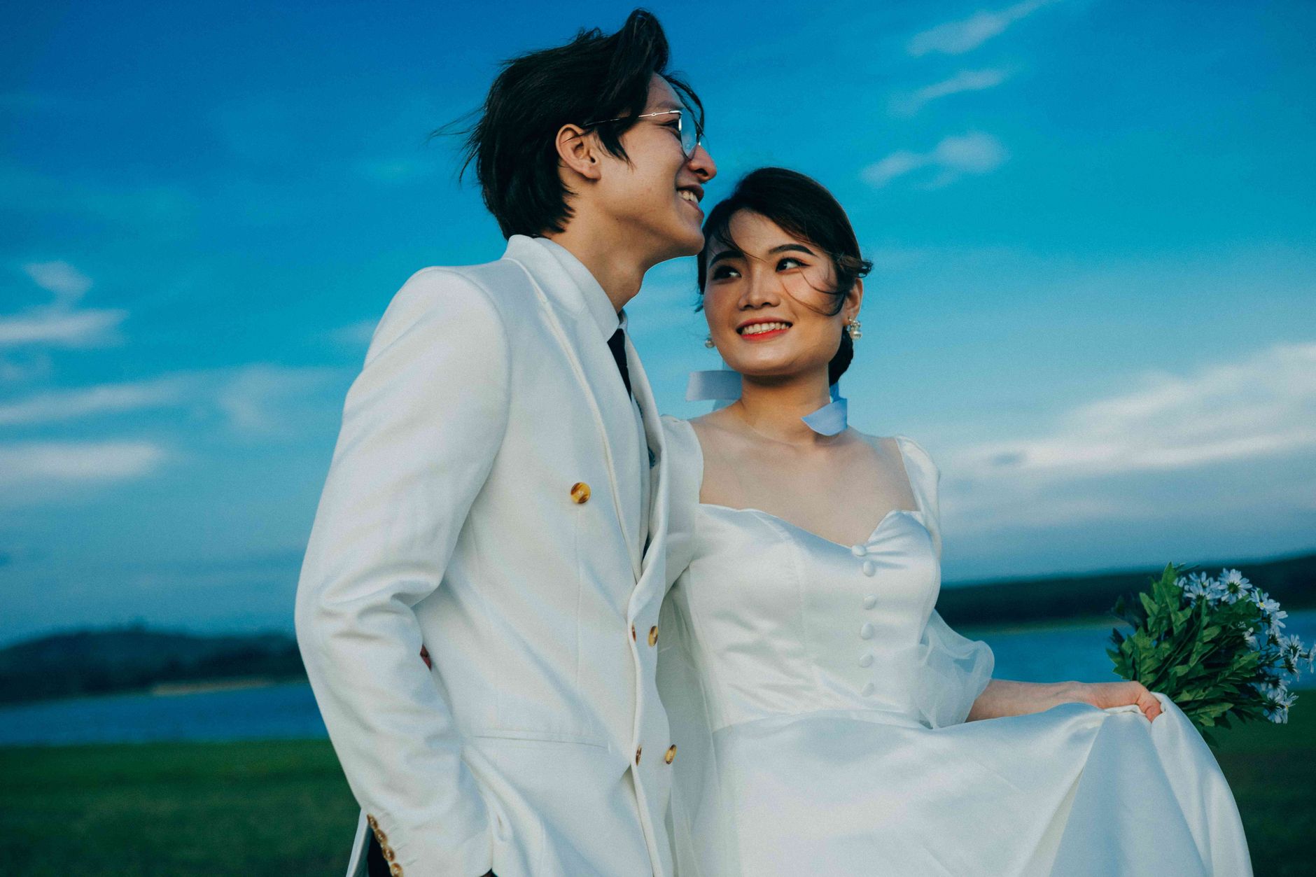 Happy newlywed couple outdoors in elegant wedding attire, radiating joy and romance against a vibrant sky.