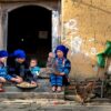A family sits outside a traditional village home, engaging in daily activities.