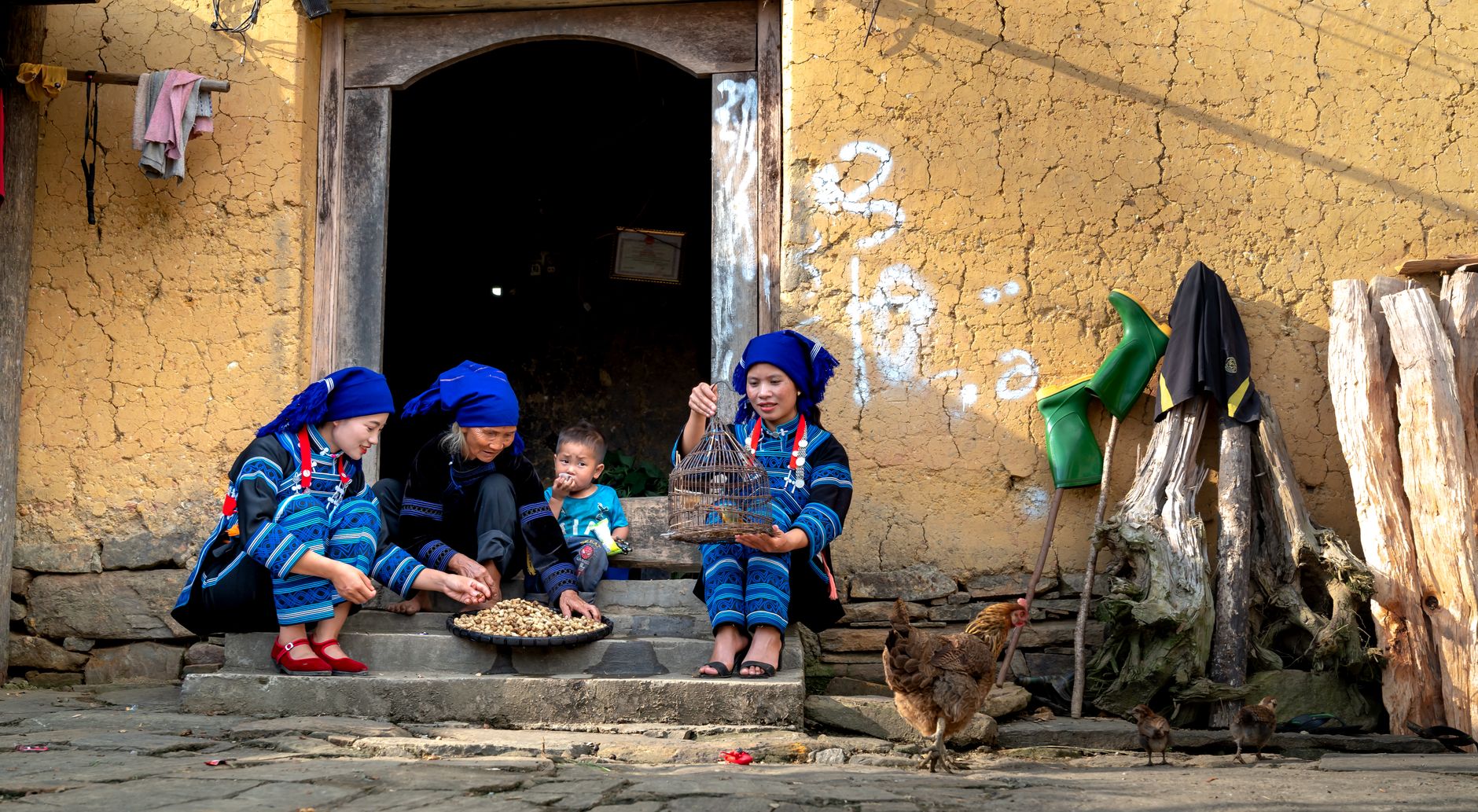 A family sits outside a traditional village home, engaging in daily activities.
