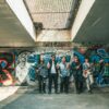 A group of young musicians with guitars standing in front of colorful graffiti in an urban setting.
