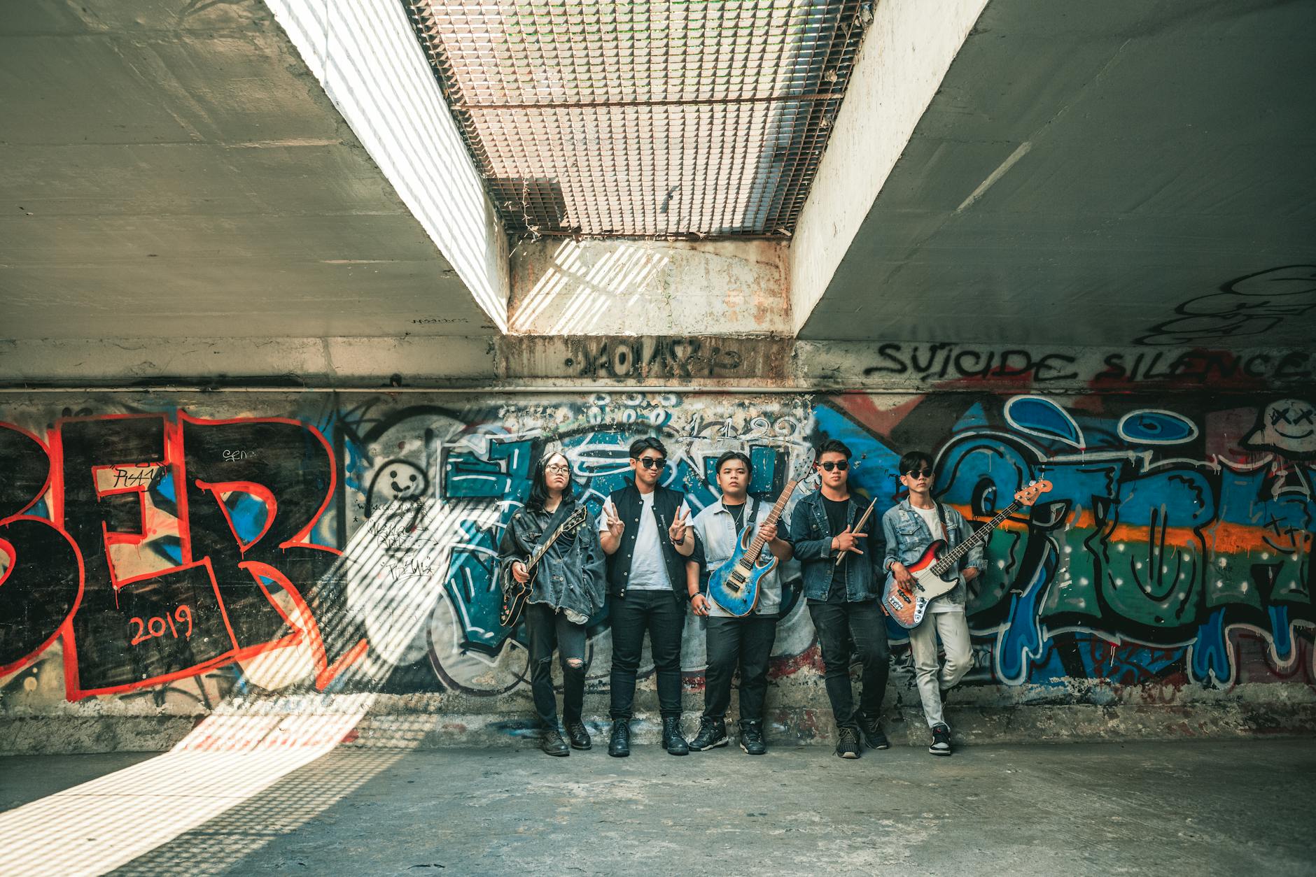 A group of young musicians with guitars standing in front of colorful graffiti in an urban setting.