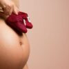 Close-up of a pregnant woman holding red baby shoes on her belly, symbolizing anticipation and love.