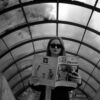Black and white photo of a woman reading a magazine on an urban escalator, wearing sunglasses.