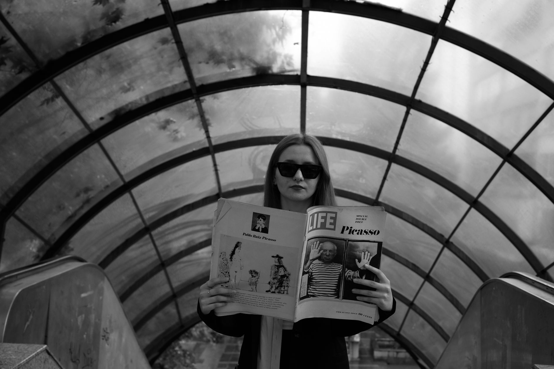 Black and white photo of a woman reading a magazine on an urban escalator, wearing sunglasses.