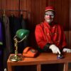 A clown bellhop in costume stands at a wooden desk with colorful attire, ready to entertain.