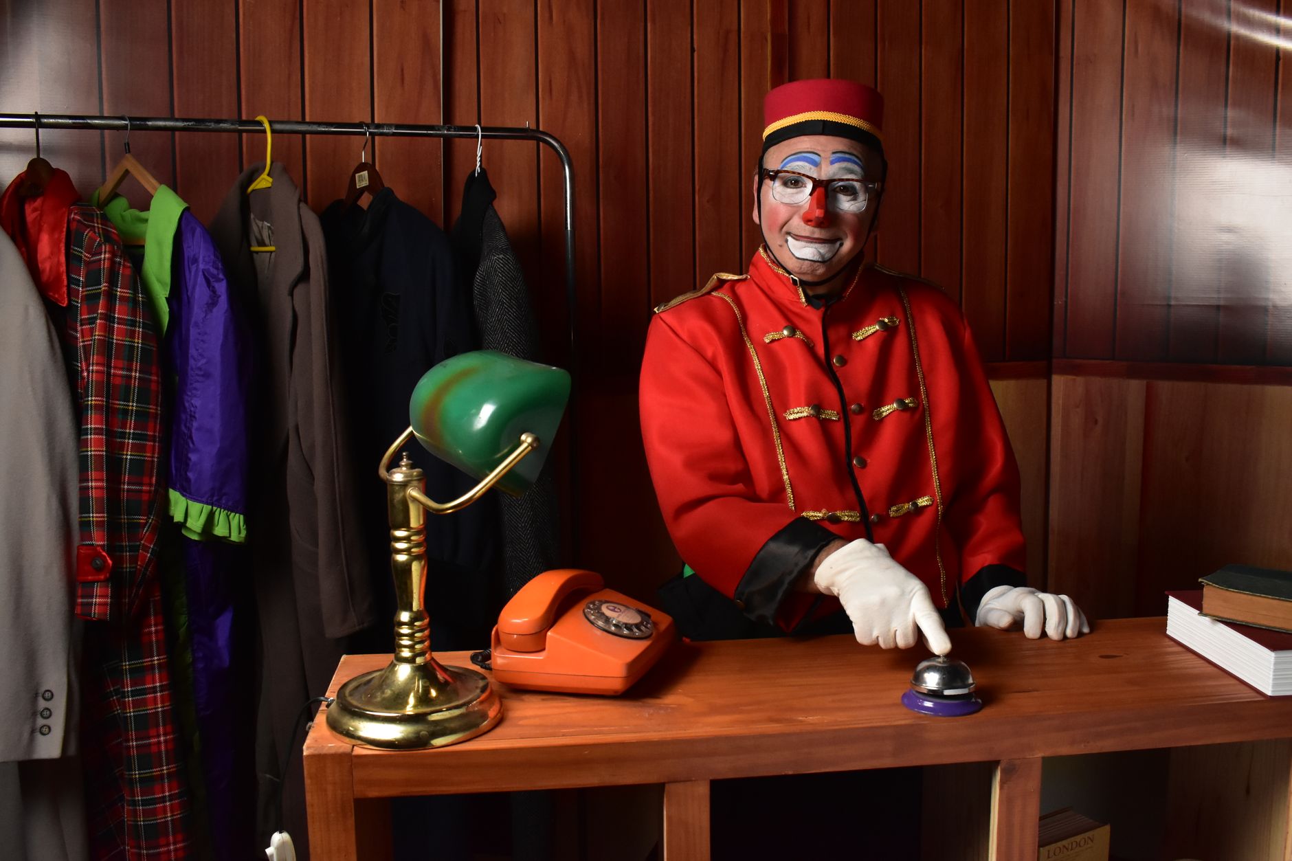 A clown bellhop in costume stands at a wooden desk with colorful attire, ready to entertain.