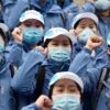 Group of healthcare workers wearing masks and blue uniforms holding fists up, showing unity and strength.