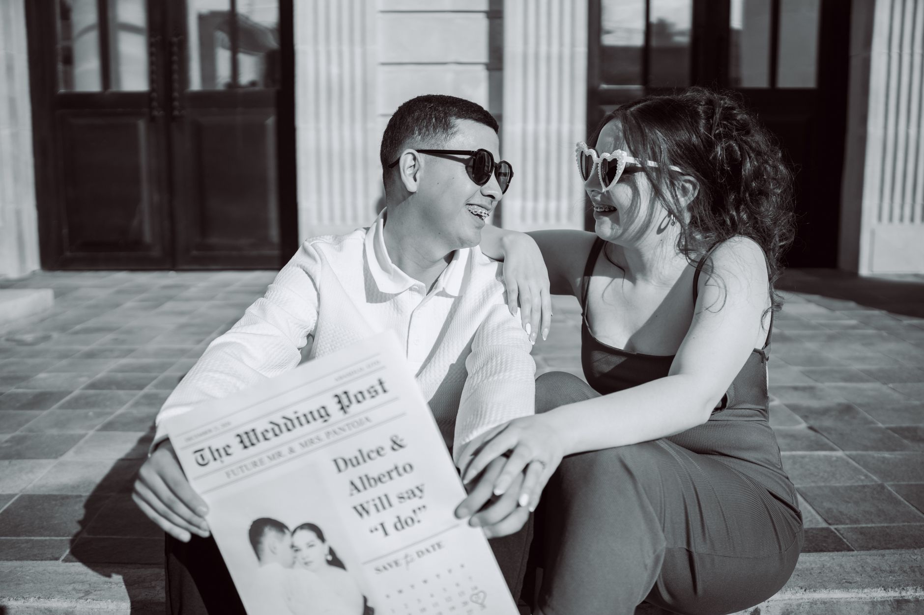 A happy couple sharing their engagement news with sunglasses and newspaper in hand.