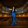 A woman in a stunning blue dress performs on a grand theater stage with arms raised.