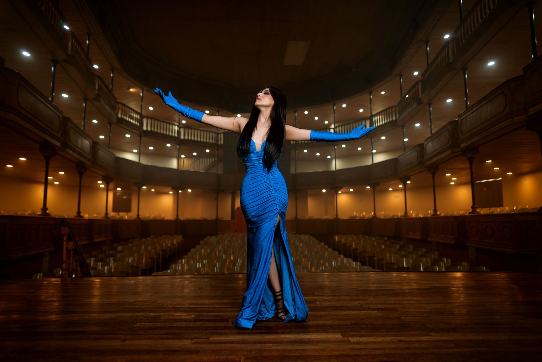 A woman in a stunning blue dress performs on a grand theater stage with arms raised.
