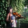 A cosplayer dressed in vibrant attire plays the violin amidst lush greenery, showcasing artistic expression.