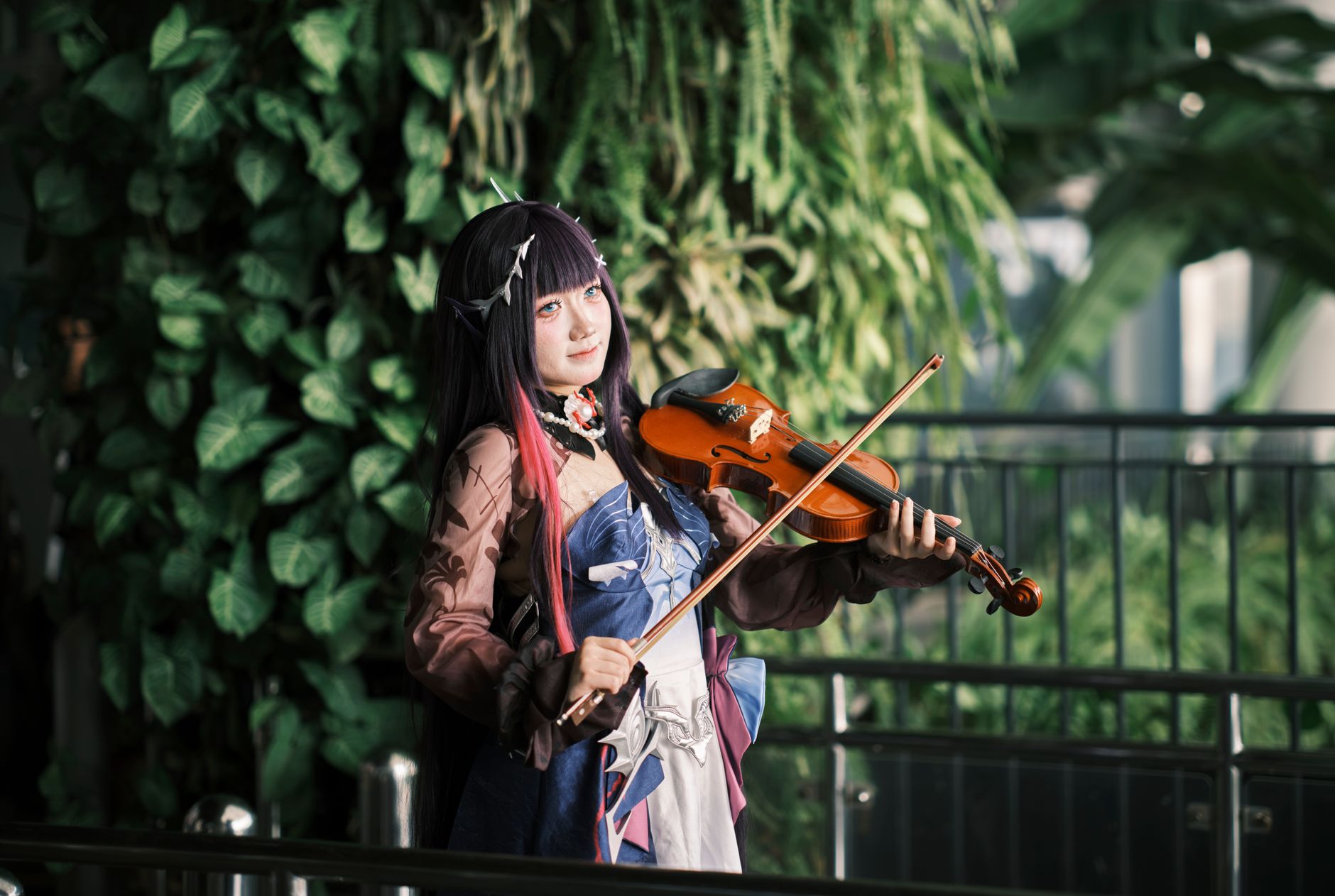 A cosplayer dressed in vibrant attire plays the violin amidst lush greenery, showcasing artistic expression.