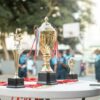 A golden trophy displayed on a table during an outdoor sports event with blurred players in the background.