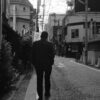 A man walks alone down a quiet street in a Japanese city, captured in black and white.