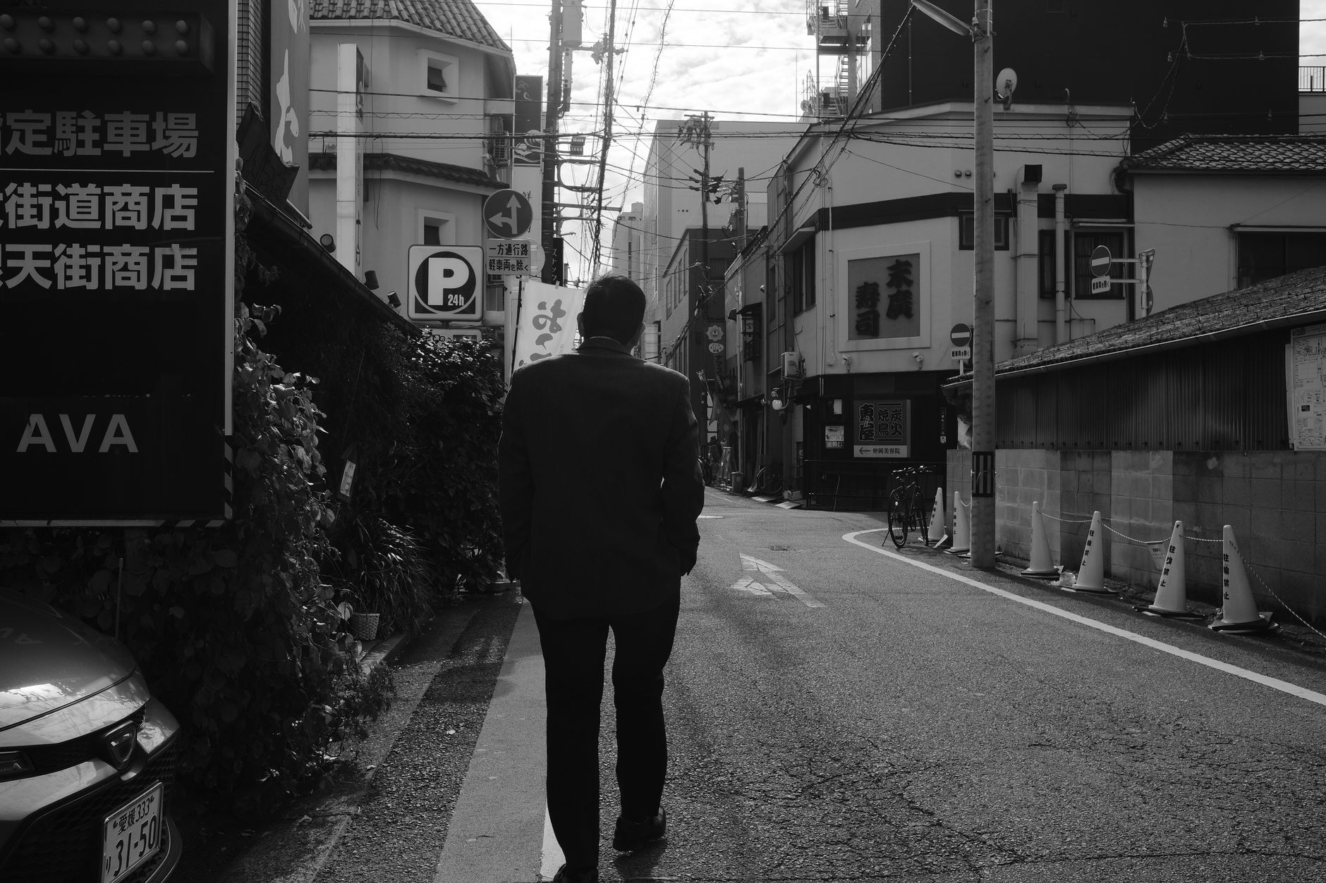 A man walks alone down a quiet street in a Japanese city, captured in black and white.