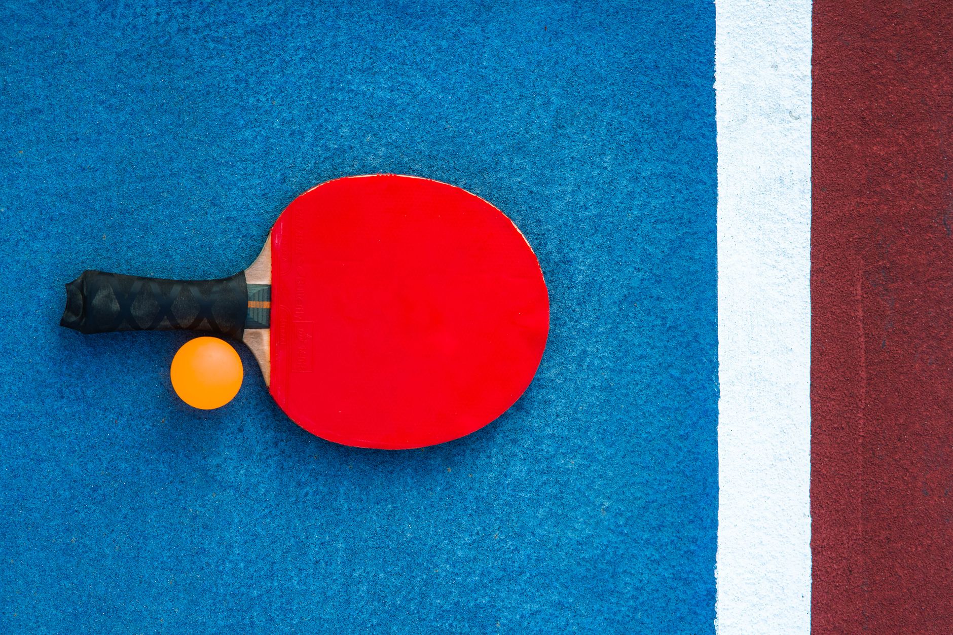 A red table tennis paddle and orange ball on a colorful court background.