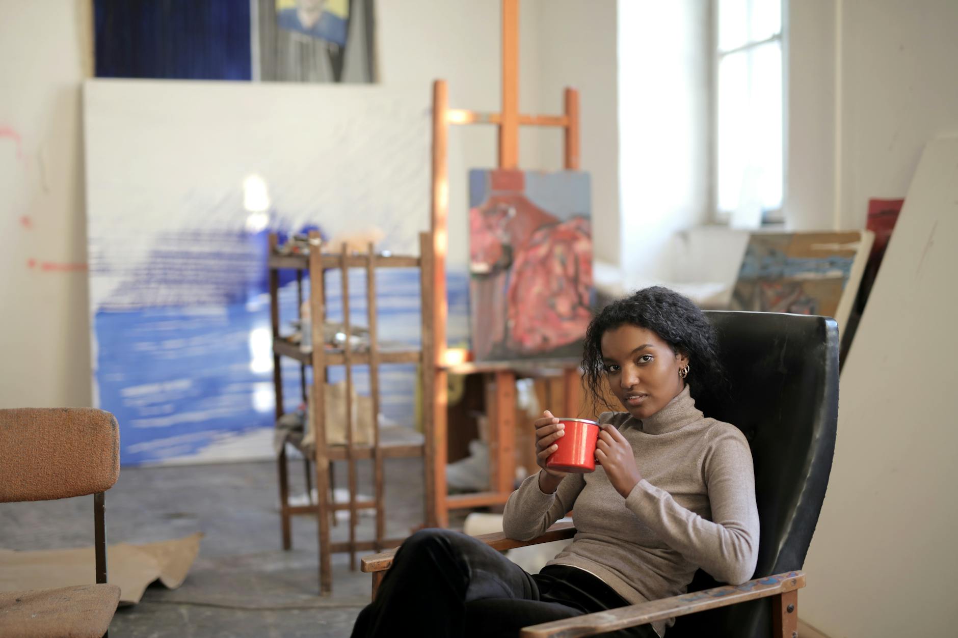 Resting black female in casual outfit sitting on comfortable old armchair and enjoying hot drink while chilling in creative art workspace