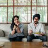 Pensive Indian male having meal on sofa while woman female in casual clothes sitting near and switching TV channels