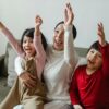 Cheerful Asian mom and kids watching sport on TV at home and screaming happily while cheering with raised arms