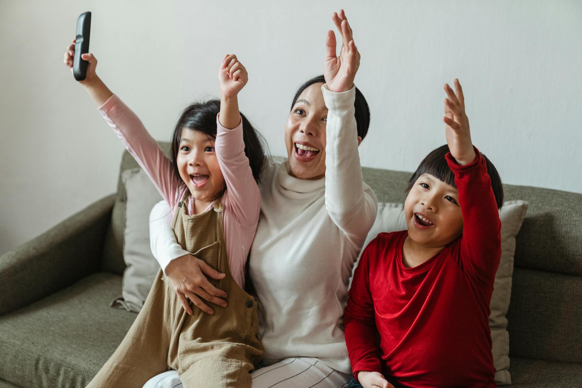 Cheerful Asian mom and kids watching sport on TV at home and screaming happily while cheering with raised arms