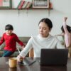 Asian mother working online using laptop and taking notes on tablet with stylus and cheerful children making noise and running behind in living room