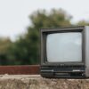 Small black old fashioned television placed on stone railing on street in city with green lush trees on blurred background
