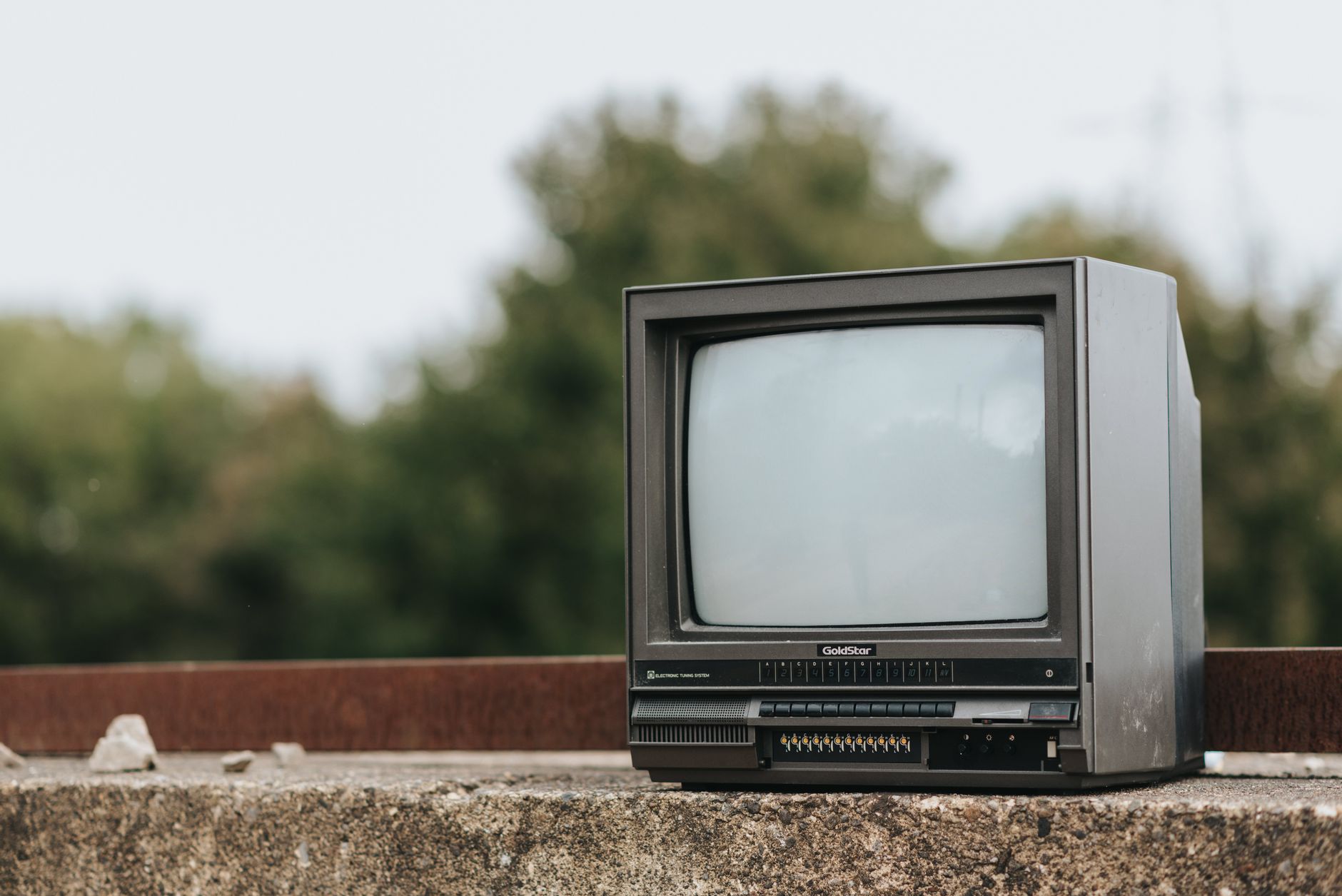 Small black old fashioned television placed on stone railing on street in city with green lush trees on blurred background