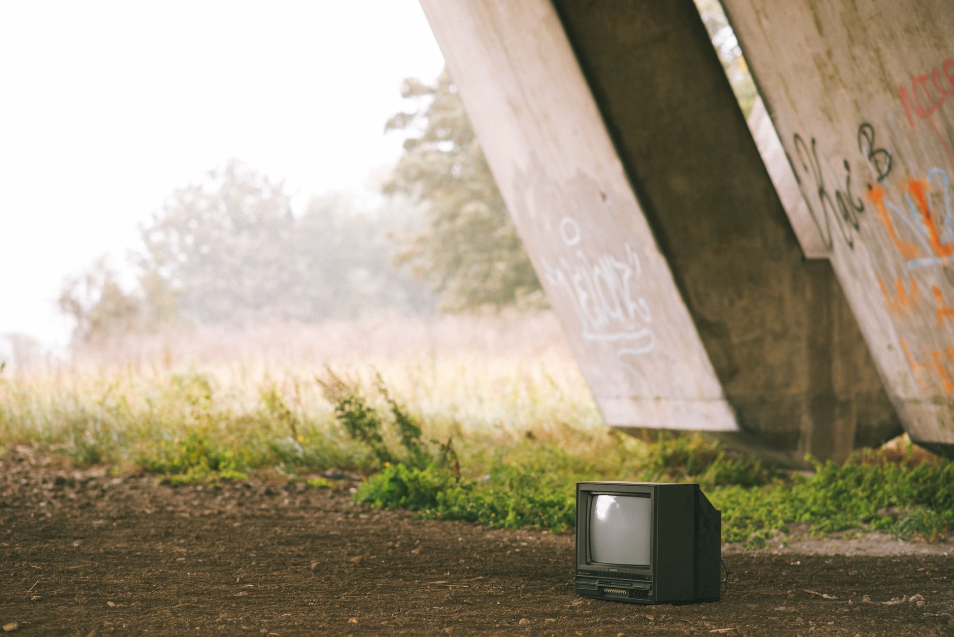 Small black old fashioned television placed on ground near concrete construction with graffiti and grassy terrain with trees on foggy background