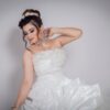 Female model dressed in bridal gown with makeup and jewelry touching neck gently standing against gray background