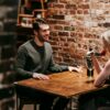 Young couple in elegant clothes sitting in light cafeteria at wooden table near brick wall and having romantic dinner with red rose and wine near glasses