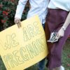 A couple holds a yellow sign saying 'We Are Pregnant' outdoors with red flowers in the background.