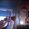 Musician playing guitar and singing on stage while audience cheers with drinks indoors.