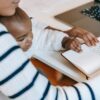 Ethnic baby with mother wearing casual outfit while sitting at table with opened notebook near laptop in bright room