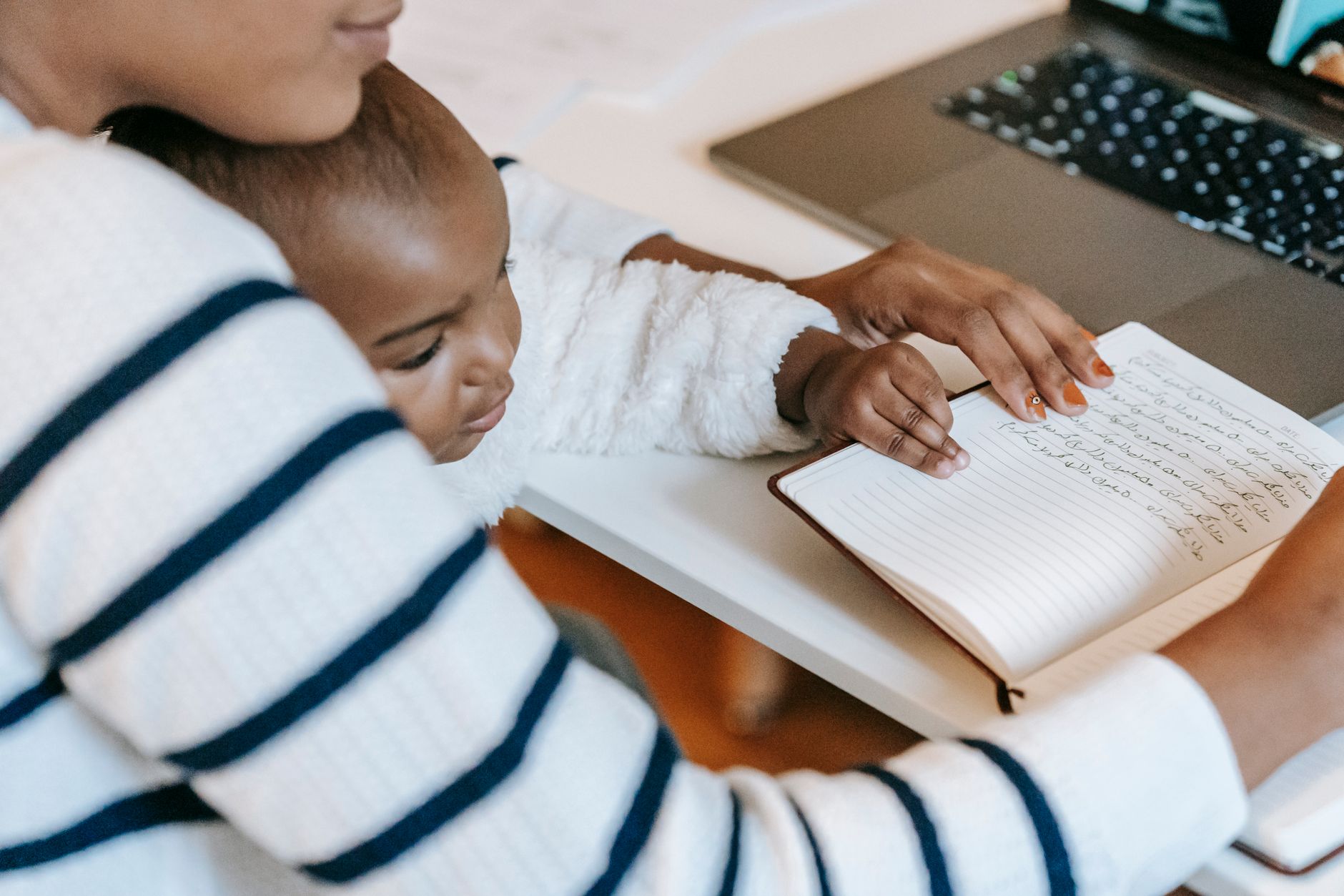 Ethnic baby with mother wearing casual outfit while sitting at table with opened notebook near laptop in bright room