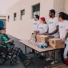 Volunteers assist a senior adult in a wheelchair by distributing aid packages outdoors.