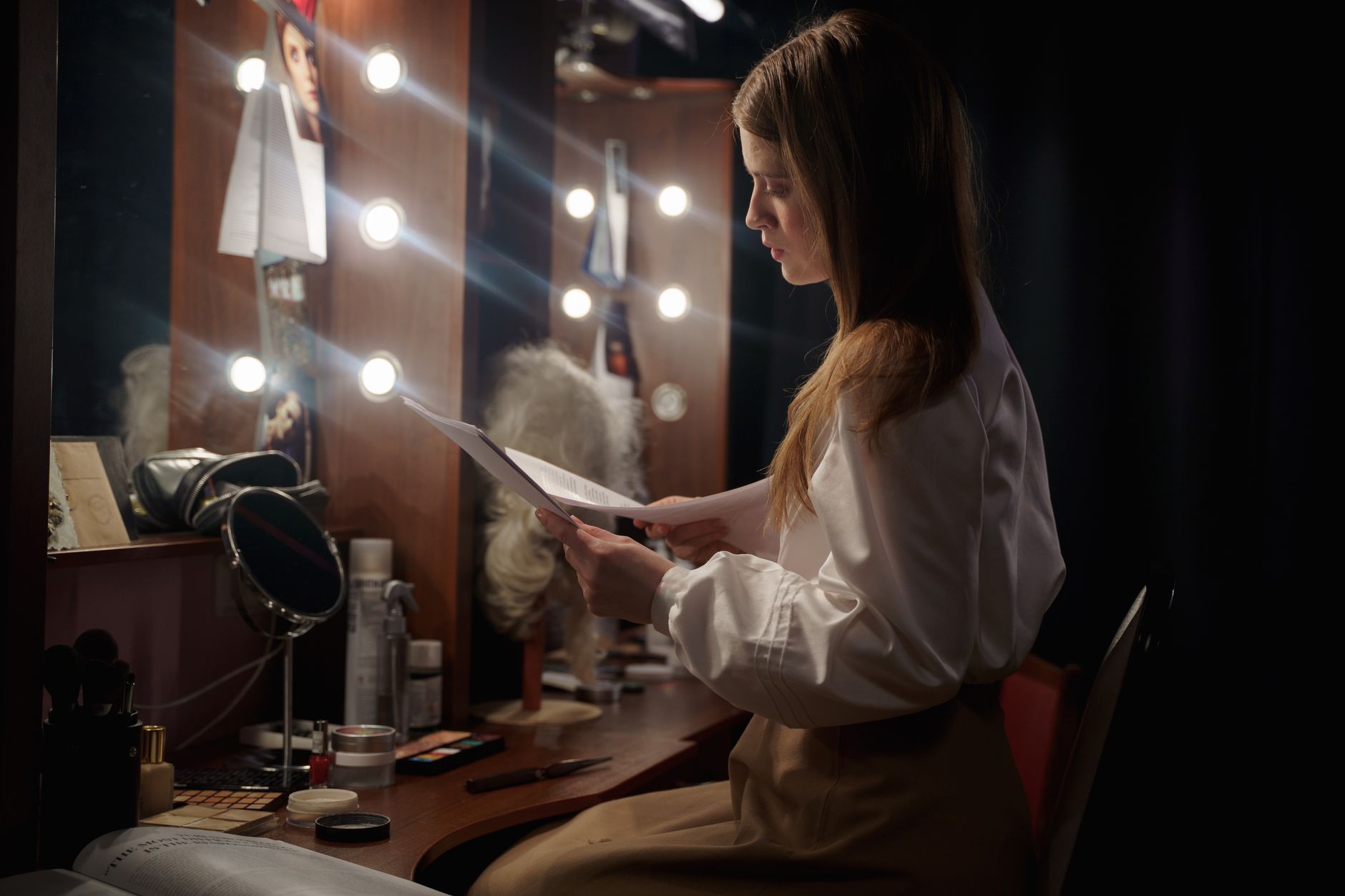 A woman in a dressing room reading a script, surrounded by makeup and lights.