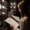 Actress reading script in a dressing room, surrounded by makeup and lights.