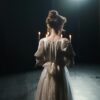 An actress in a white dress stands backstage in dim lighting, enhancing a classic theatrical atmosphere.