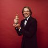 Smiling man in a tuxedo holding a trophy against a red background, celebrating success.