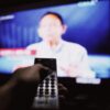 Crop anonymous person with remote controller switching channels while watching TV on blurred background in living room with dim light