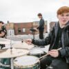 A young drummer energetically performs with a band on a rooftop, showcasing musical passion.