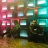 Two individuals in a cyberpunk setting with neon lighting and stacked television screens.